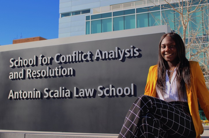 A young African American woman in a bright yellow blazer and plaid black and white dress pants sits in front of a university sign. She is smiling.